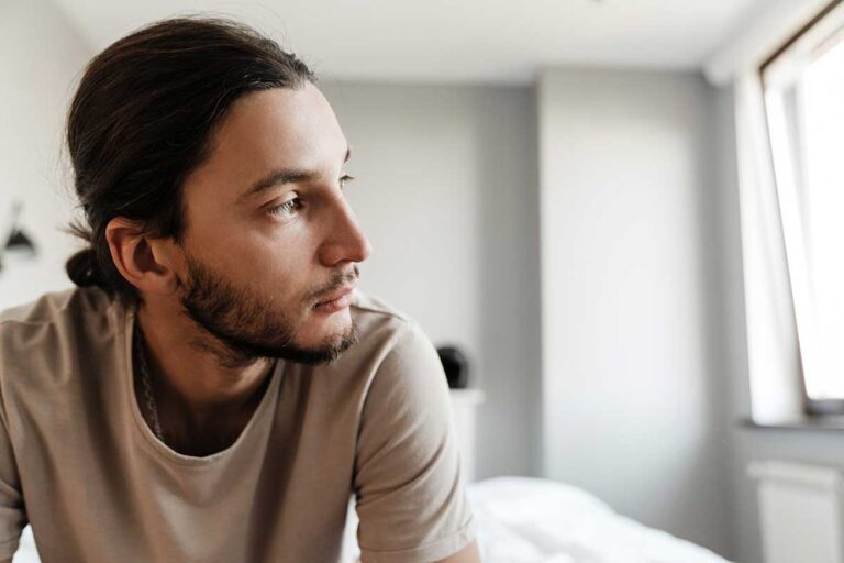 person sitting on bed looking out window while struggling with alcohol abuse and depression